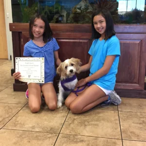 Two girls kneel on a tile floor with a dog between them; one girl holds a certificate.