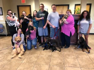 A group of people pose indoors with several dogs of various breeds, some sitting and some being held.
