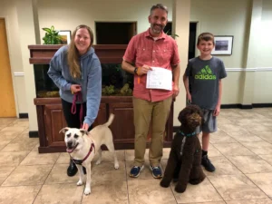 Three people and two dogs pose indoors; one man holds a certificate and both dogs are on leashes.