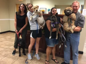 Four people standing indoors, each holding or standing with a dog, posing for a group photo in a lobby area.