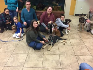 A group of people pose indoors with several dogs on leashes, sitting and kneeling on a tiled floor.