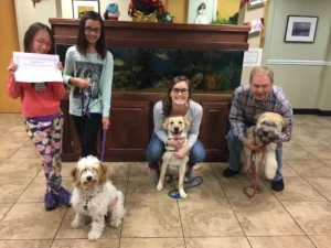 Three people and a child pose indoors with three dogs in front of a large aquarium; the child holds a certificate.