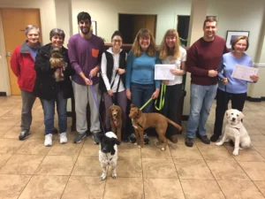 Eight people stand indoors with five dogs; two people hold certificates, and everyone is smiling at the camera.