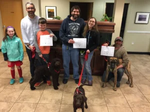 Three adults and three children hold certificates, standing with three dogs on leashes in an indoor setting.