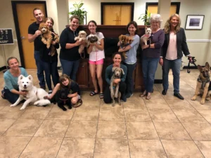 A group of people pose indoors with their dogs, sitting and standing on a tiled floor in front of wooden cabinets.