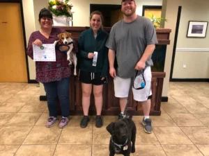 Three people and two dogs stand indoors; one person holds a certificate, another holds a ribbon, and the dogs are on leashes.