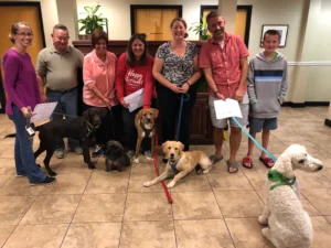 Seven people stand indoors with five dogs on leashes, holding certificates, in a tiled room with plants and wooden counters.