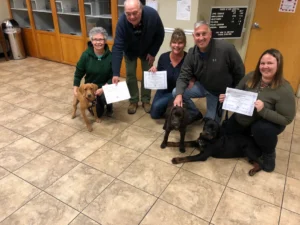 Five adults kneel indoors with three dogs, each holding certificates, suggesting a dog training class graduation.