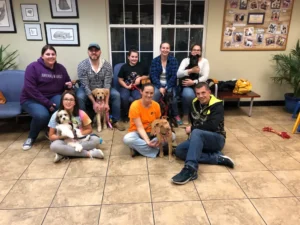 A group of people sit indoors with their dogs, posing for a group photo in a room with tile floors and wall decorations.
