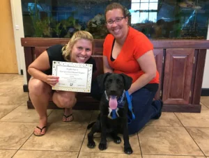 Two women pose with a black dog wearing a blue bandana; one woman holds a certificate.