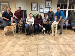 A group of people and their dogs pose together indoors in a waiting room with tiled floors and framed art on the walls.