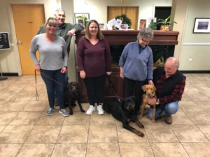 Five adults and three dogs pose indoors in front of a large aquarium, all looking toward the camera and smiling.