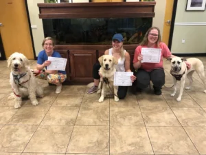 Three women kneel beside their dogs holding certificates, posing indoors in front of an aquarium.