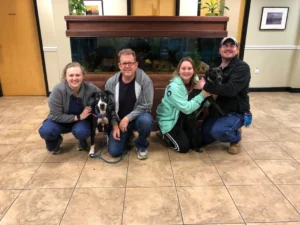 Four people kneel indoors with two black dogs in front of a large fish tank and wood-paneled wall.