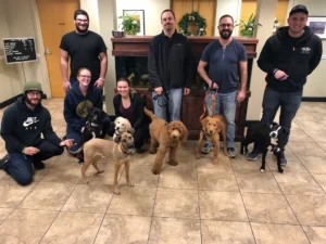 Seven people pose indoors with five dogs of various breeds on leashes in front of a wooden aquarium stand.