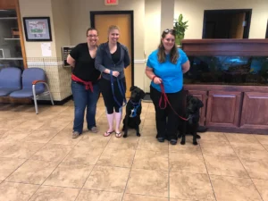 Three women stand indoors with two black dogs on leashes in a waiting area with chairs and a large fish tank.