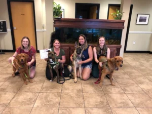 Four women kneel indoors with four dogs in front of a large fish tank; one woman holds a certificate.