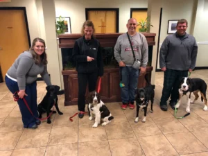 Four people stand indoors with four dogs on leashes in front of a fish tank, all facing the camera.