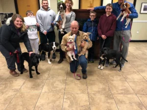 A group of people pose indoors with several dogs of various breeds, some sitting and some being held on leashes.