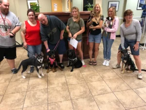 Seven people stand indoors with their dogs on leashes, posing for a group photo in a tiled room.