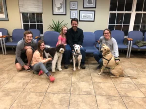 Five people and three dogs sit and kneel together in a waiting room with blue chairs and framed art on the walls.