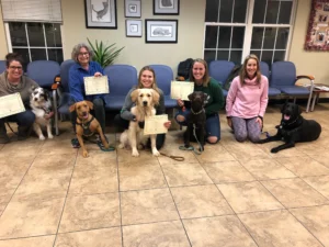 Five women sitting with their dogs, each holding a certificate, in a room with tile floors and blue chairs.