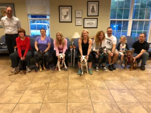 Nine people sit in a waiting room with six dogs, posing for a group photo; artwork and windows are in the background.